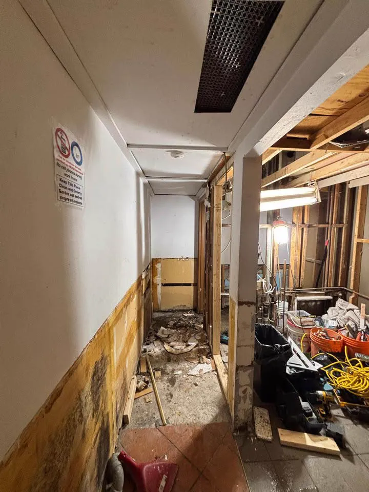 Narrow commercial corridor stripped along one side with framing exposed and debris staged