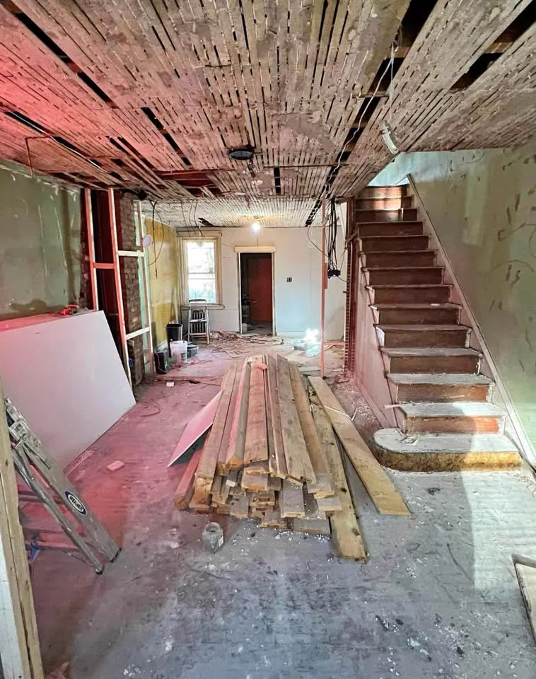 Basement stripped to the lath ceiling with the stair framing exposed and lumber stacked