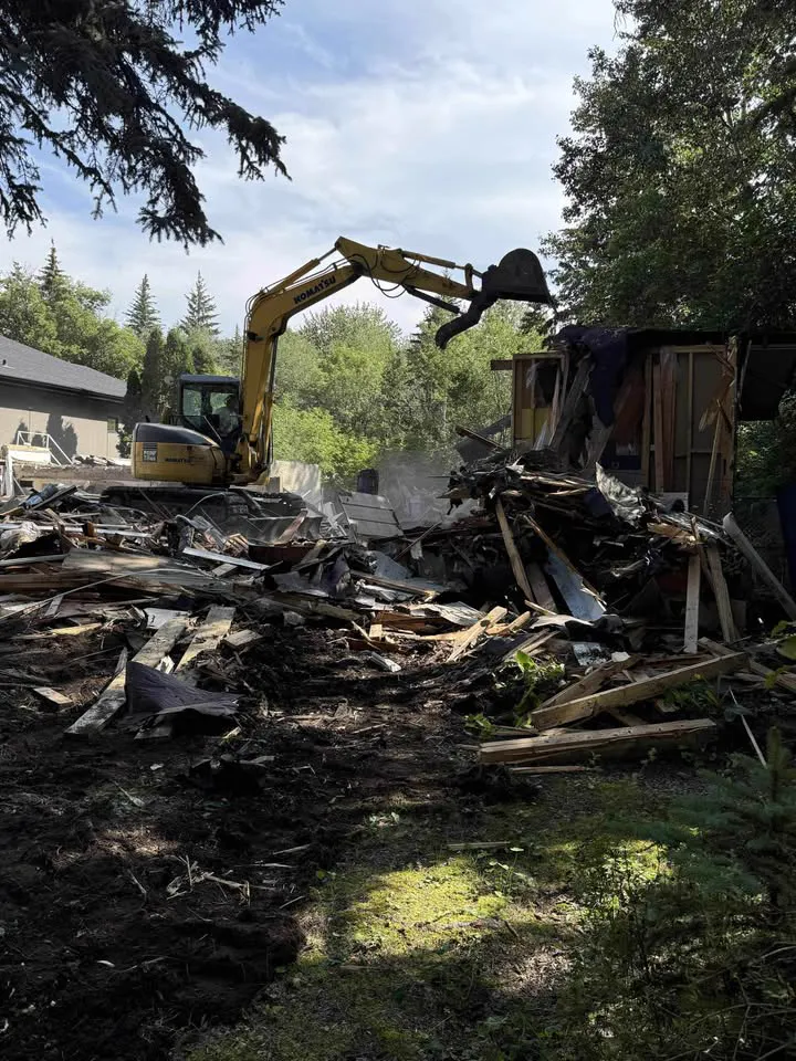 Excavator knocking down a small wood-frame building beside trees