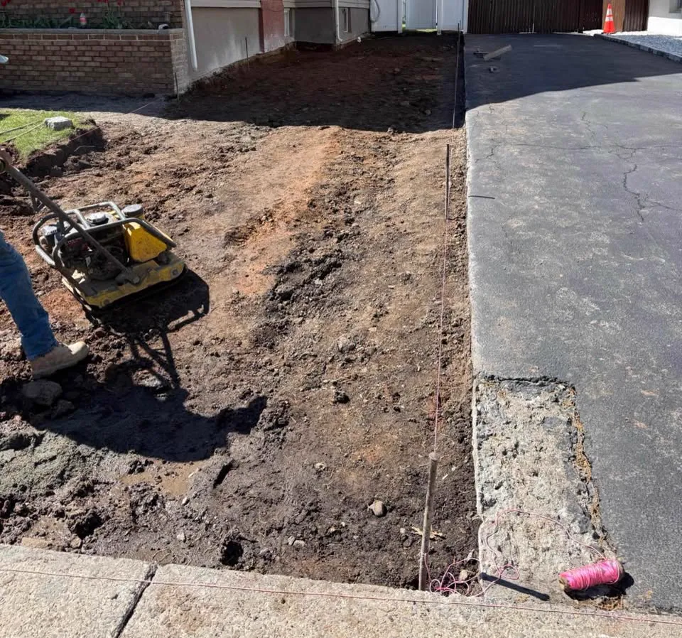 Excavated strip beside a driveway graded down to soil with a plate compactor